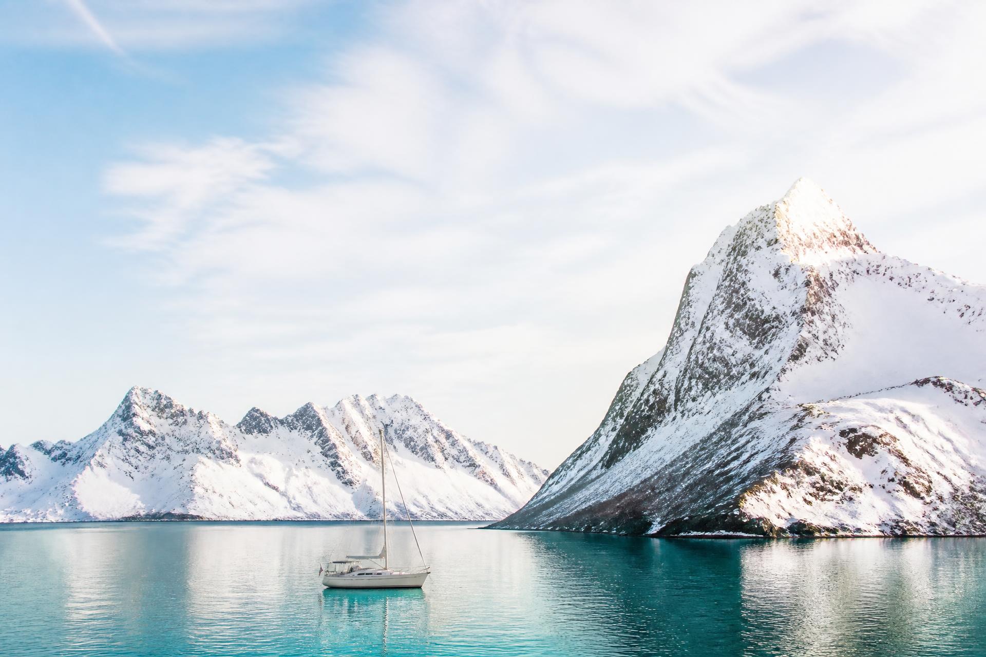 A lone sailboat anchored in a sunlit Arctic fjord beneath snow-covered peaks